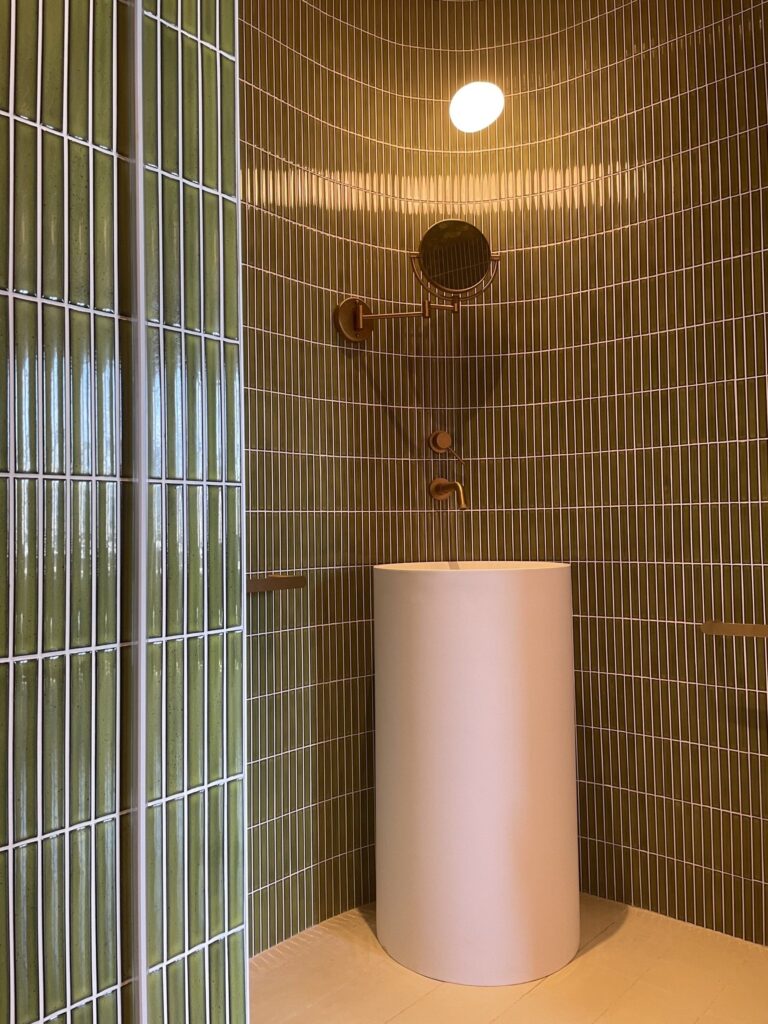 Photo+16-07-2022-+11+27+00-be490c23-1920w.jpg Modern bathroom with green vertical tiles, a round sink, and a brass faucet under a ceiling light.
