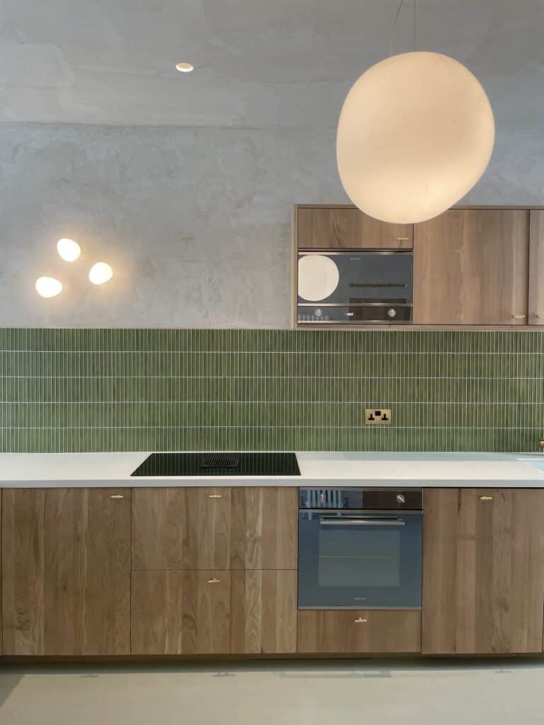Photo+16-07-2022-+12+03+10-1920w.jpg Modern kitchen with green tile backsplash, wooden cabinets, and a round hanging light.