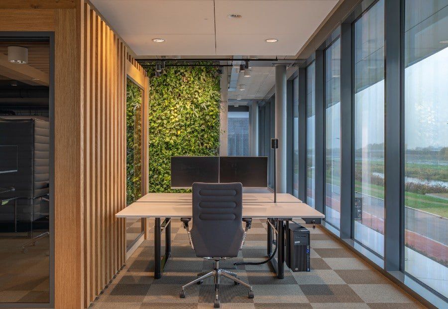 Modern office workspace with large windows, green plant wall, and a desk with a computer chair.