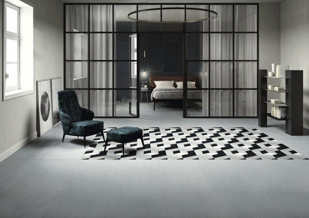 Modern bedroom with glass walls, patterned rug, dark chair, and shelf in a minimalist setting.