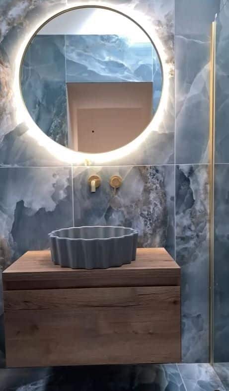 Modern bathroom with round backlit mirror, blue marble tiles, vessel sink, and wooden vanity.