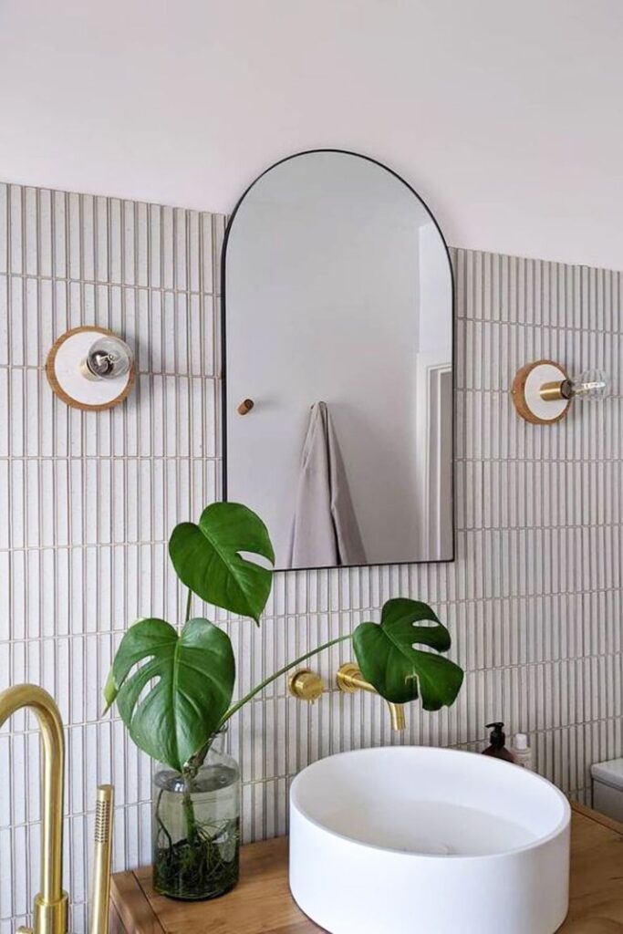 Kit+Kat+mosaic-1920w.jpg Modern bathroom with a round mirror, white sink, gold fixtures, and a vase with green leaves.