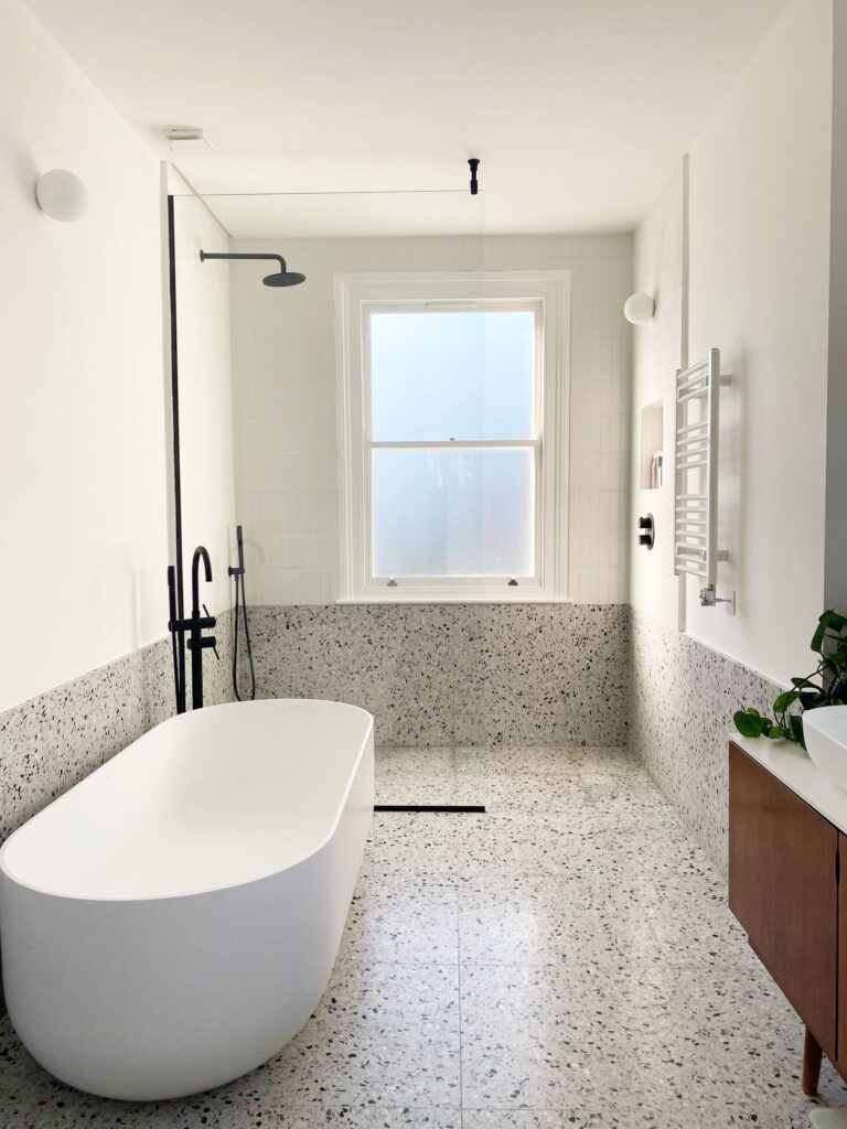 bathroom+2+terrazzo+7+2-df7d5dea-1920w.jpg Modern bathroom with terrazzo floor, white bathtub, window, and minimal decor.