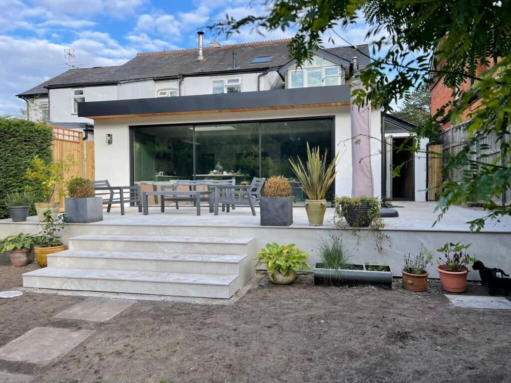 outdoor+shot+5-69ca6dc7-1920w.jpg Modern house with a large glass patio door, outdoor dining area, and potted plants on a raised terrace.