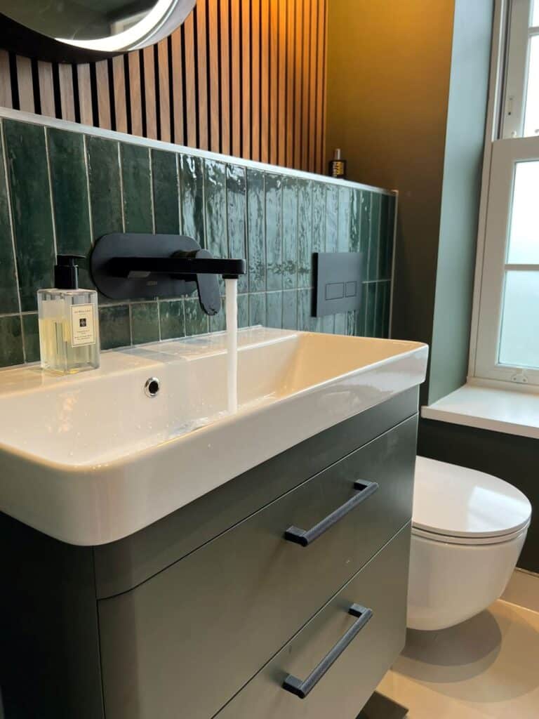 Modern bathroom with green tiles, white sink, black faucet, and a window in the background.