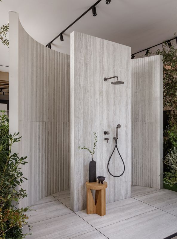 Modern outdoor shower with a wooden stool and plants surrounding the area.