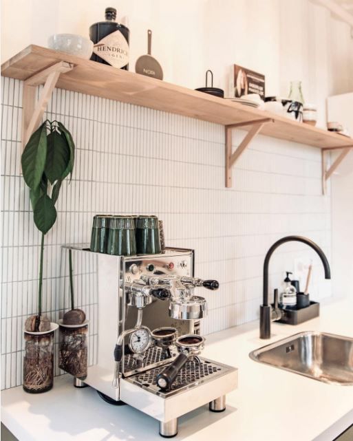 Espresso machine and cups on a white kitchen counter with a shelf and sink nearby.