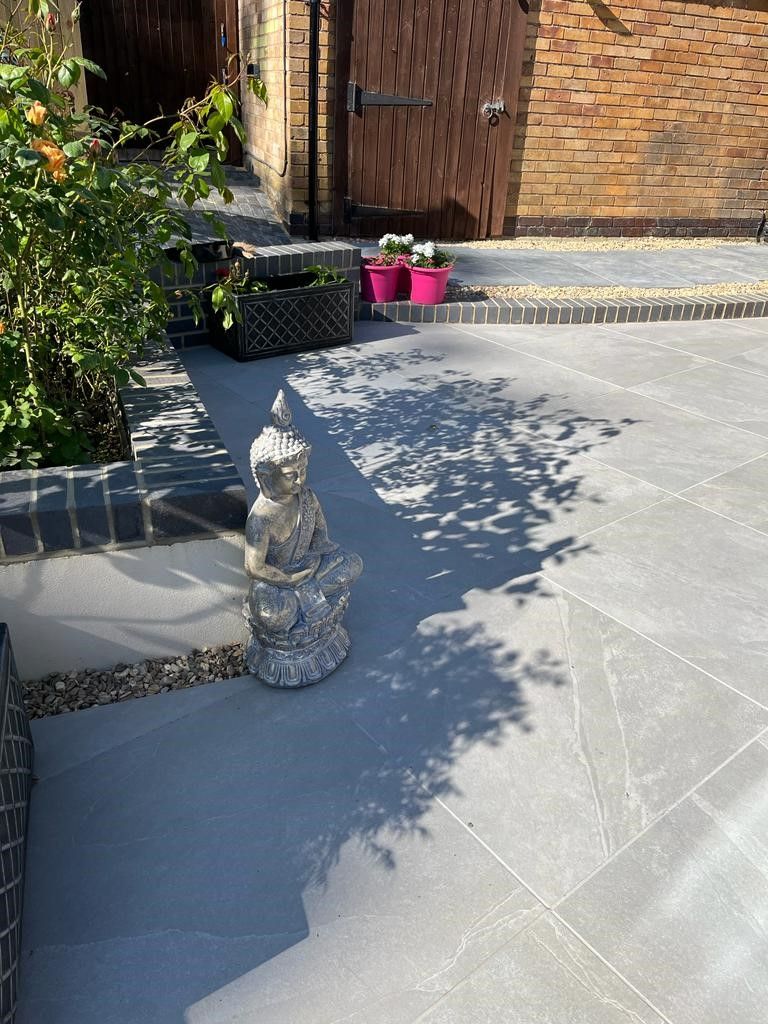 2-2c381450-1920w.jpg Stone Buddha statue on a gray patio with plants, flowers, and a brick wall in the background.