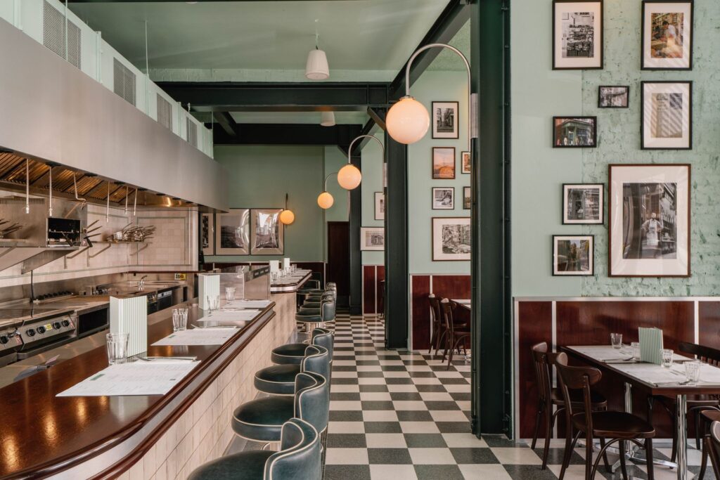 Adam+Firman_Lina+Stores_South+Kensington_Regraded-25-a285f420-1920w.jpg A retro-style restaurant with checkered floors, green walls, bar stools, and framed pictures.