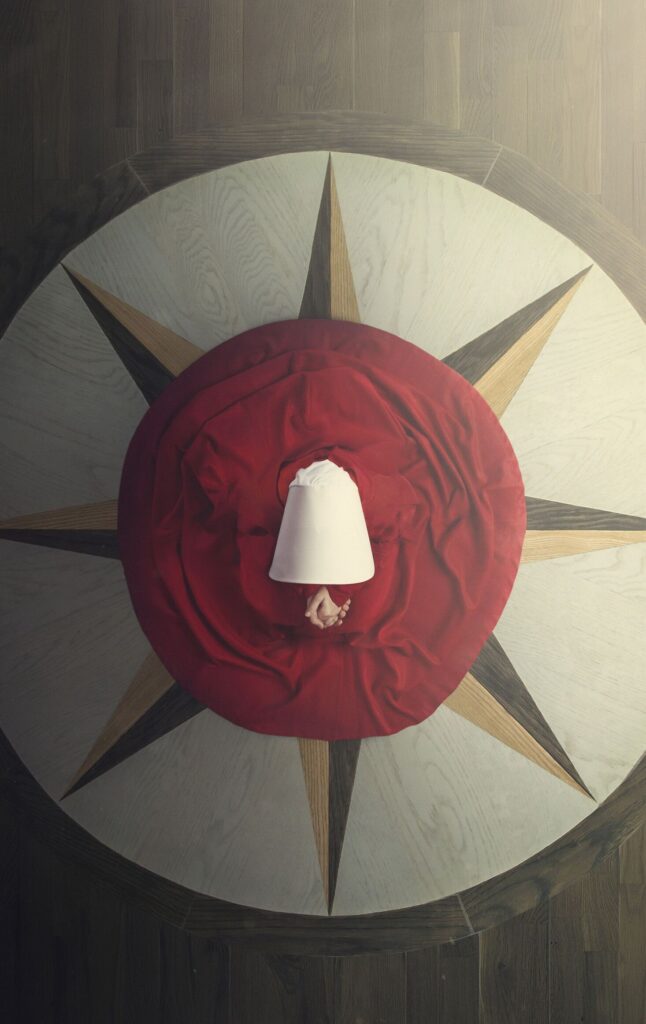 The+Handmaid-s+Tale+Exploring+the+Colonial+Revival+Interiors+of+the+Waterford+Mansion-1920w.jpg Woman in a red dress and white bonnet lying on a star-patterned floor, seen from above.
