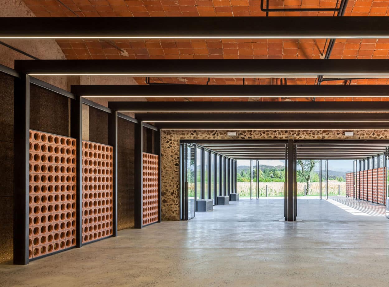 Open modern interior with concrete floor, terracotta blocks, exposed beams, and large glass windows.