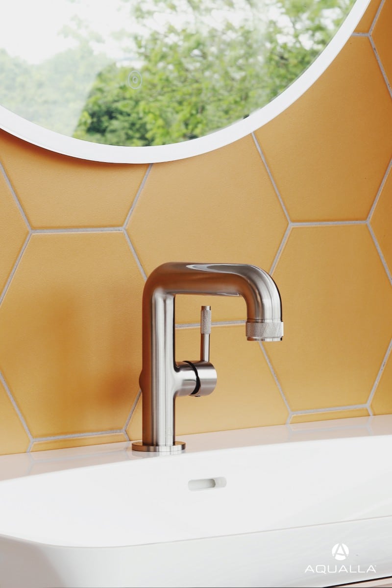 Modern silver tap on a white sink with yellow hexagon tiles and a round mirror in the background.