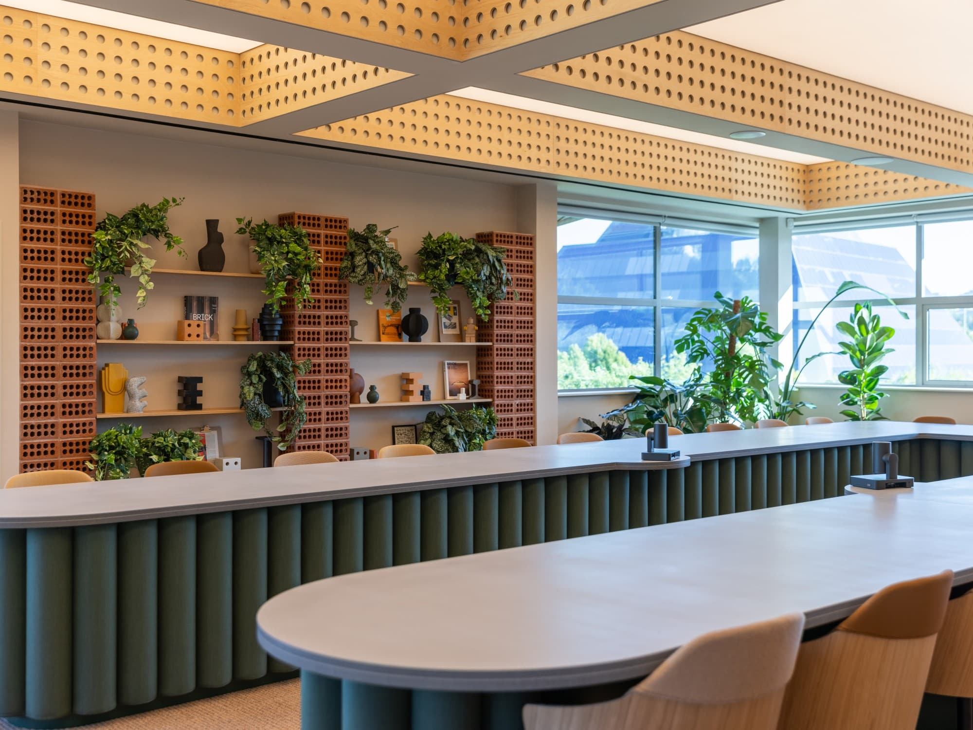 Modern office meeting room with long tables, wooden chairs, plants, and shelves with decor items.
