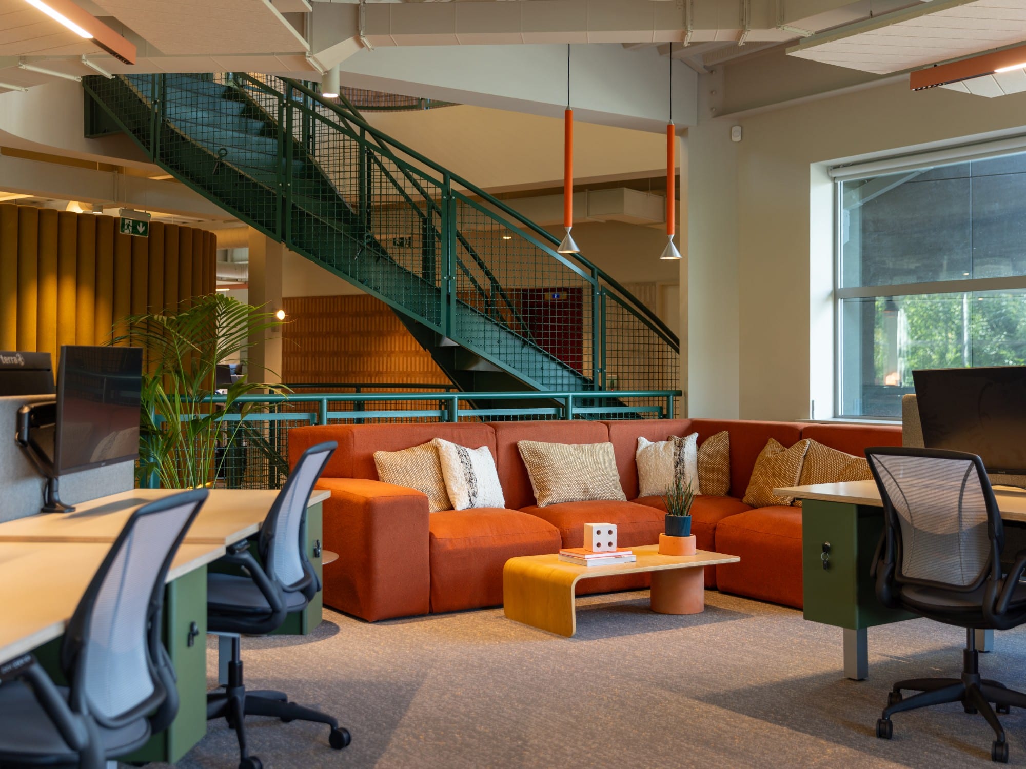 Modern office space with desks, a large orange sofa, a coffee table, and a staircase in the background.