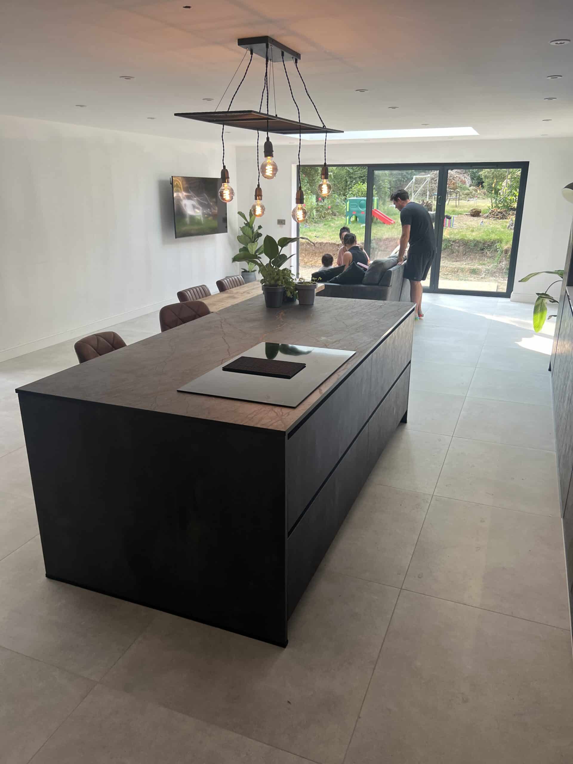 Modern kitchen with a large island, pendant lights, and people near glass doors in the background.