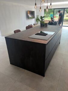 Large kitchen island with a cooktop and potted plants, modern chairs, and people in the background.