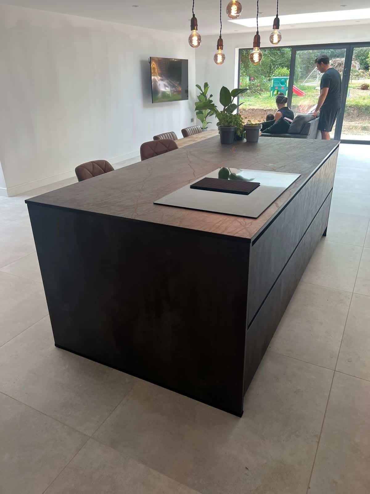Large kitchen island with a cooktop and potted plants, modern chairs, and people in the background.