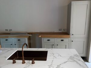 White kitchen with marble countertop, bronze sink, and light cabinets with wooden countertops.