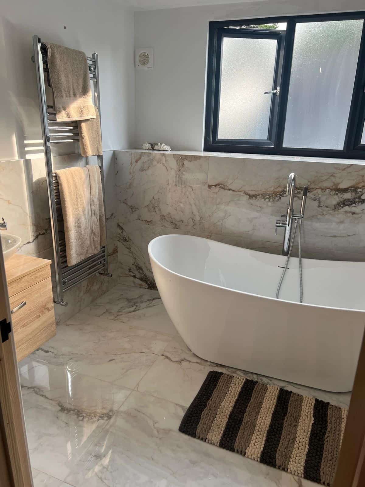 Modern bathroom with a freestanding tub, towel rack, striped rug, and large window.