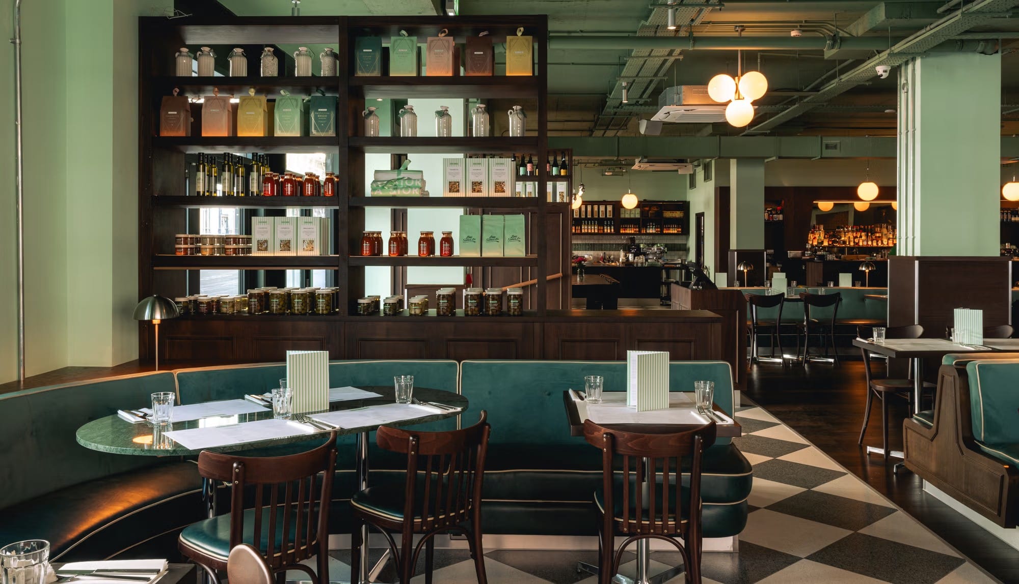 A modern restaurant interior with green booths, wooden chairs, and shelves of jars and books.