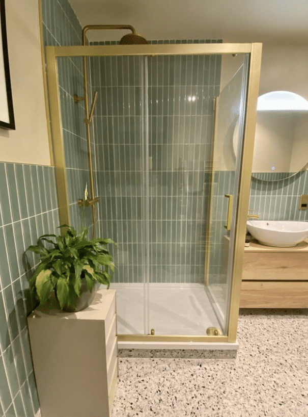 Glass shower with gold frame, green wall tiles, a white sink, and a green plant on a small stand.