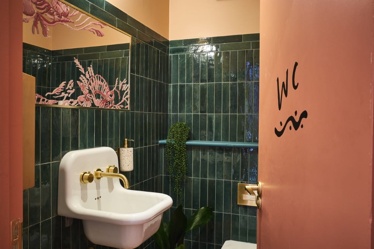 Small bathroom with green tiles, a white sink, pink door marked "WC," and a decorative mirror.