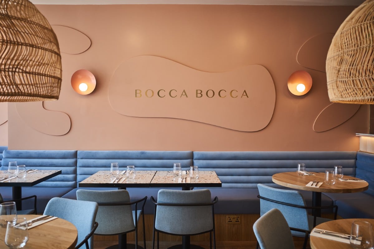 Modern restaurant interior with blue seating, wooden tables, and a sign reading "Bocca Bocca" on the wall.
