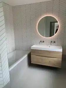 Modern bathroom with chevron wall tiles, a round backlit mirror, and a wooden vanity.