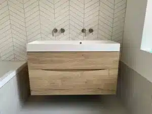 Wood vanity with white sink in front of a chevron tile bathroom wall.