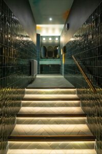 Lit stairs with green tile walls leading to a modern bathroom with three mirrors and sinks.