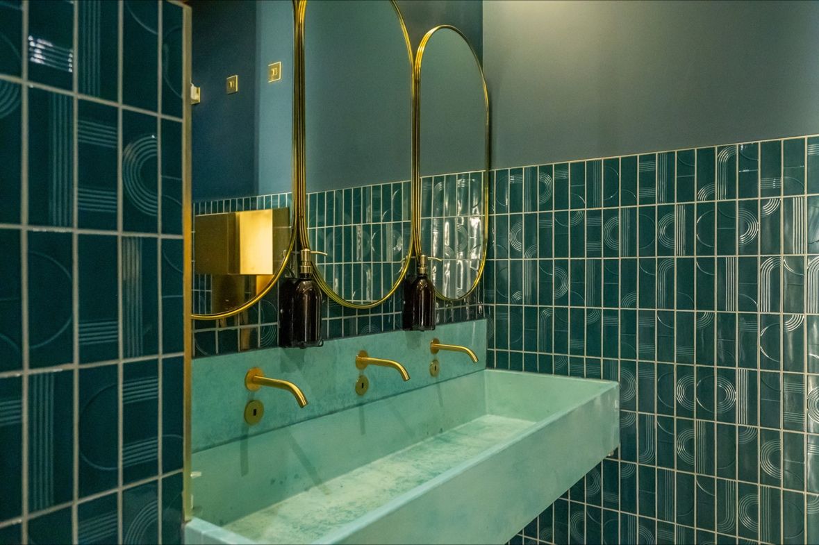 Modern bathroom with teal tiles, a light blue trough sink, gold faucets, and round mirrors.