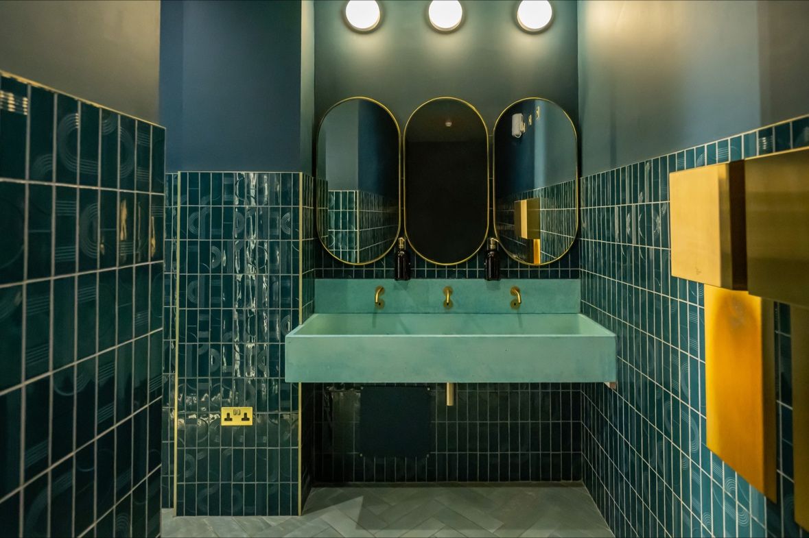 Modern bathroom with teal tiles, a long sink, three mirrors, and gold fixtures under bright lights.