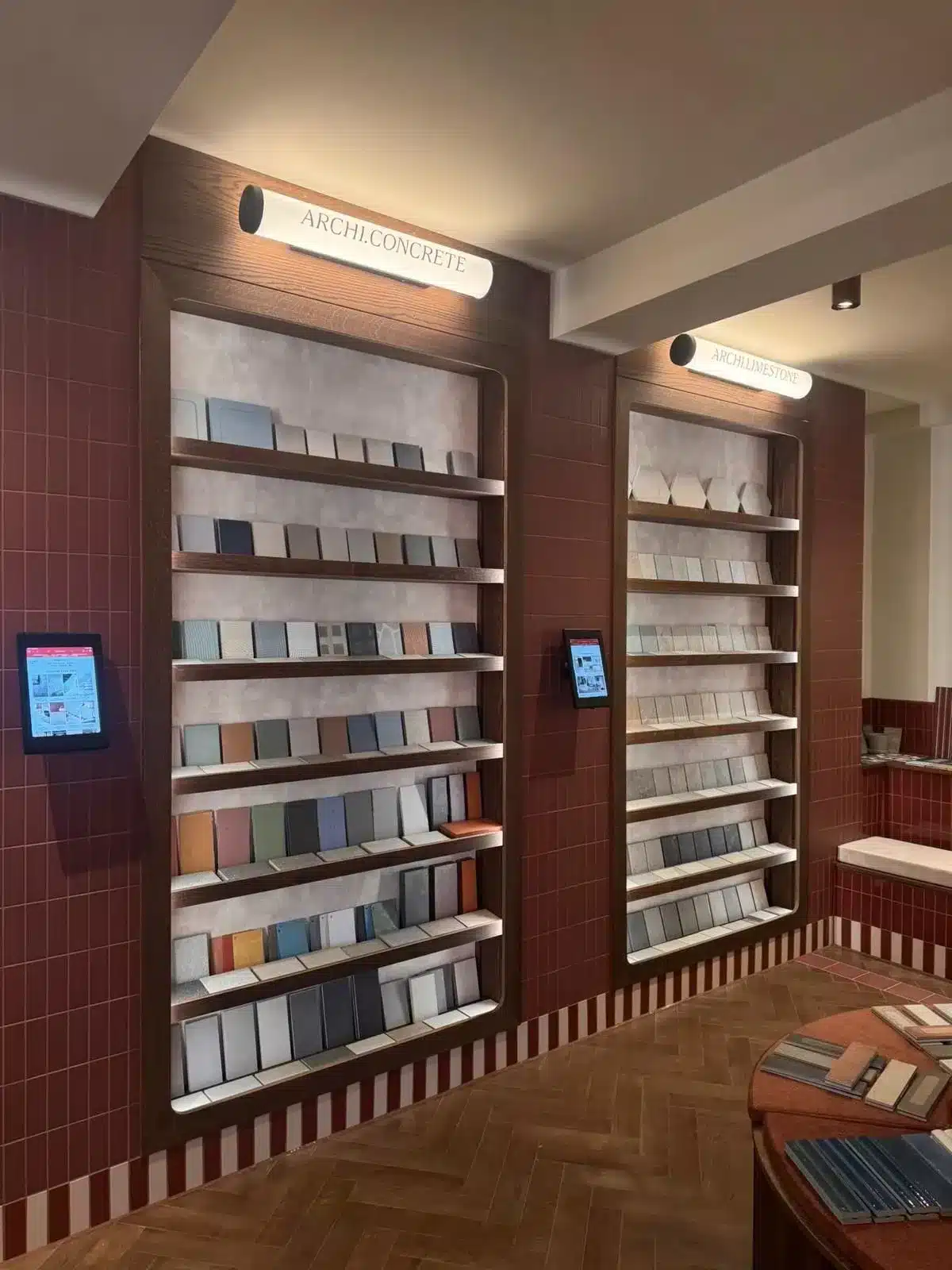 Tile showroom interior with sample tiles displayed on shelves and digital screens on a red-tiled wall.