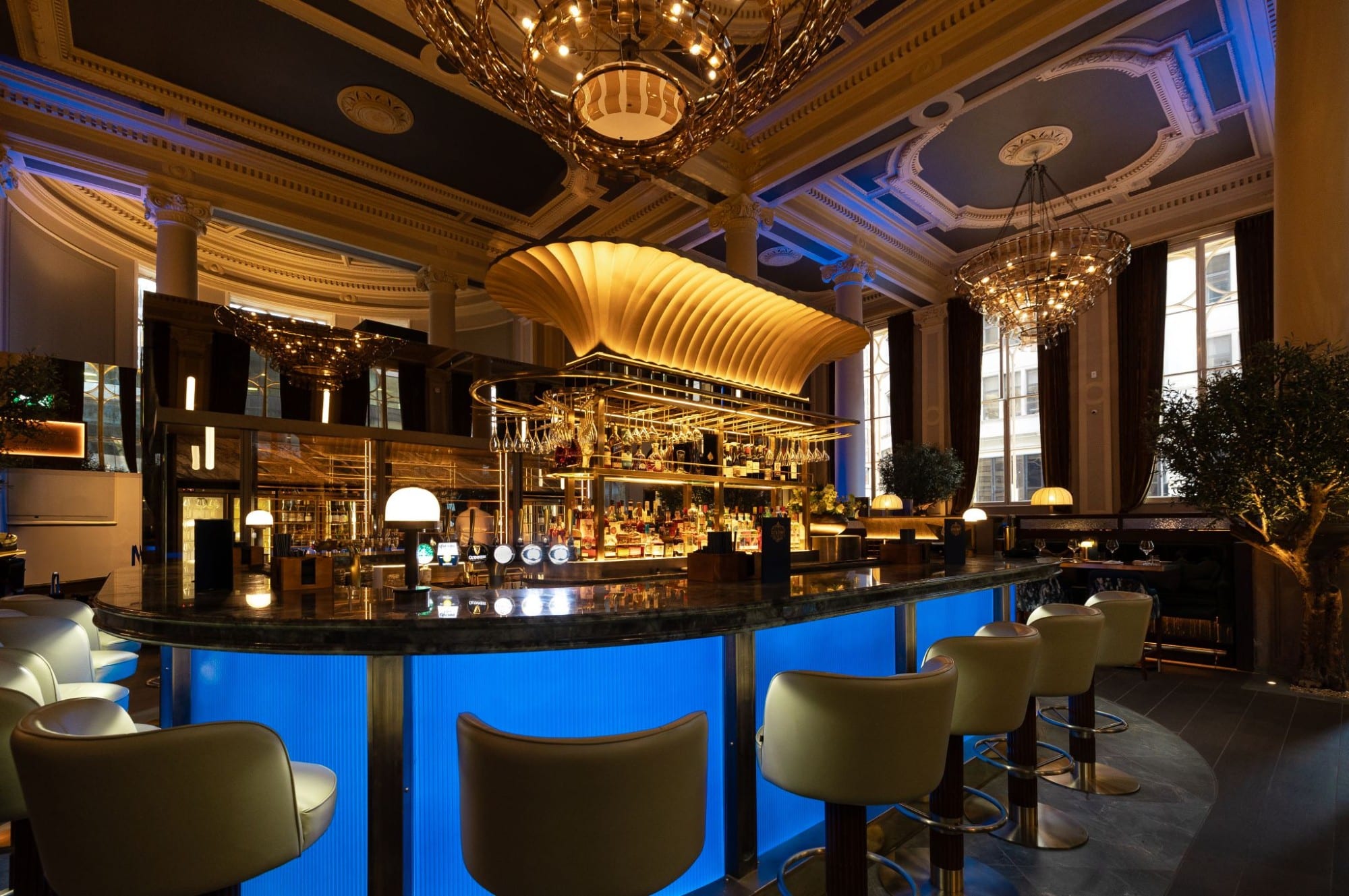 Elegant bar with glowing blue counter, white chairs, chandeliers, and high ceilings.