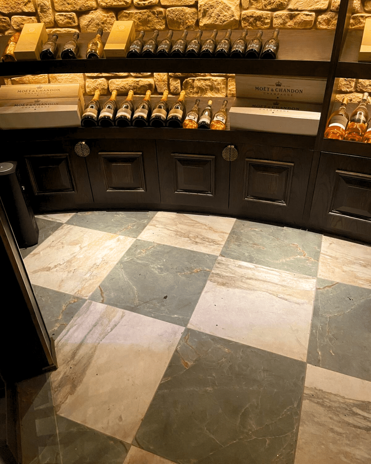 Checkerboard marble floor in a wine cellar, with bottles on wooden shelves against a stone wall.