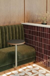 Green velvet bench, small round table, maroon tiled wall, and patterned white floor tiles.