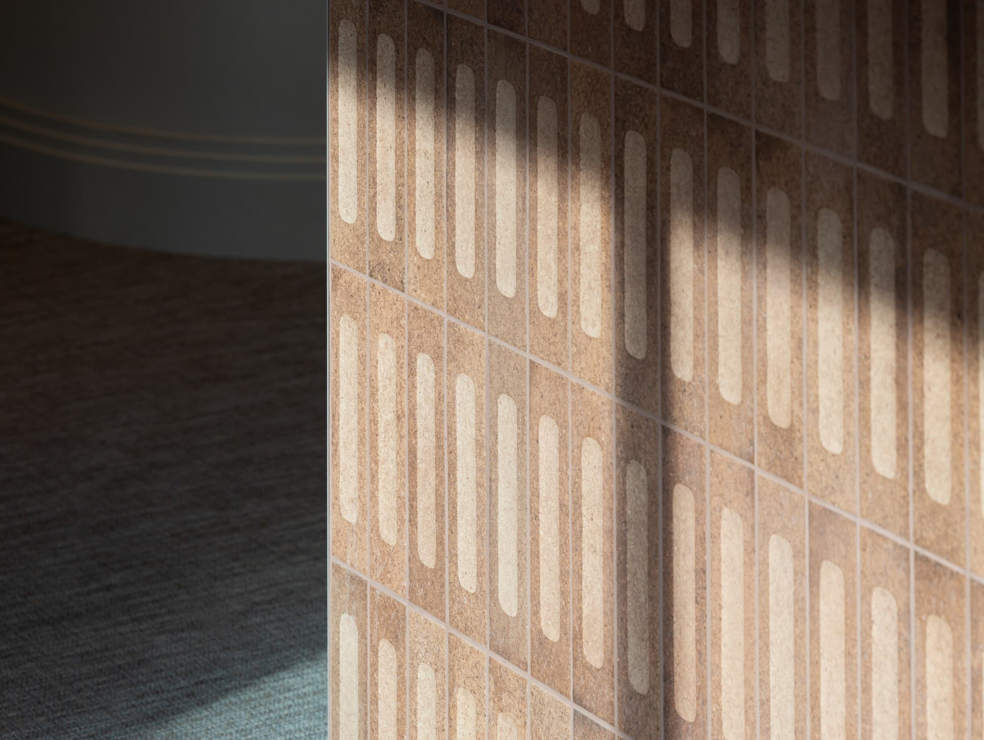 Sunlight casts a shadow on a brown tiled wall with a geometric pattern.