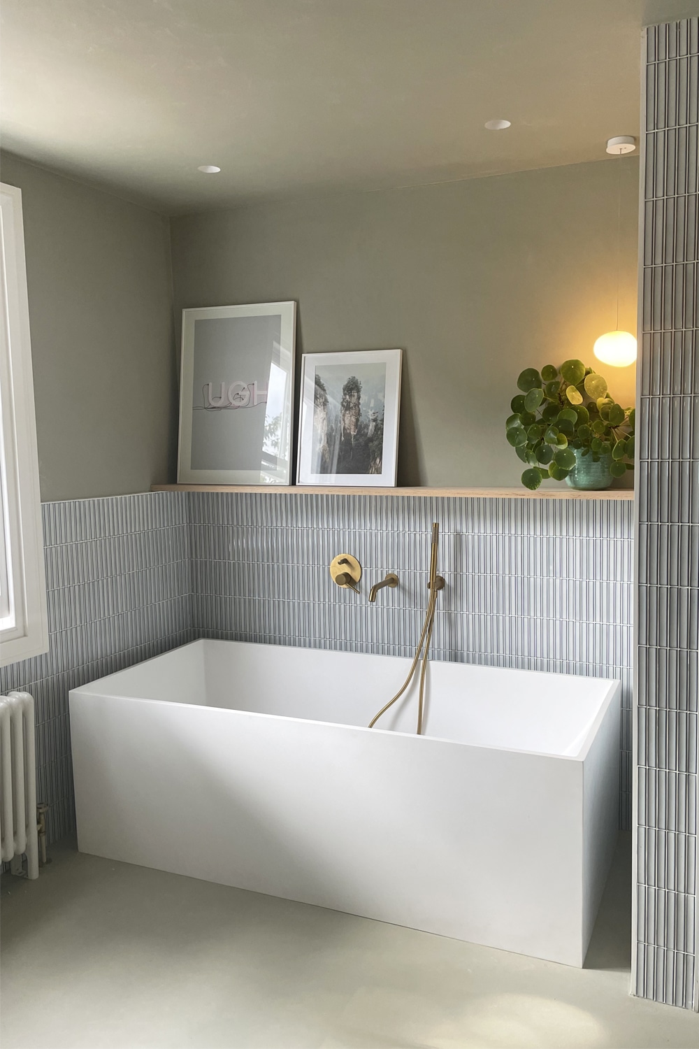 Modern bathroom with a white bathtub, grey tile, framed art, and a plant on the shelf.