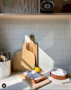 Cutting boards, a lemon, and two cakes on a kitchen counter with sunlight and a tiled backsplash.