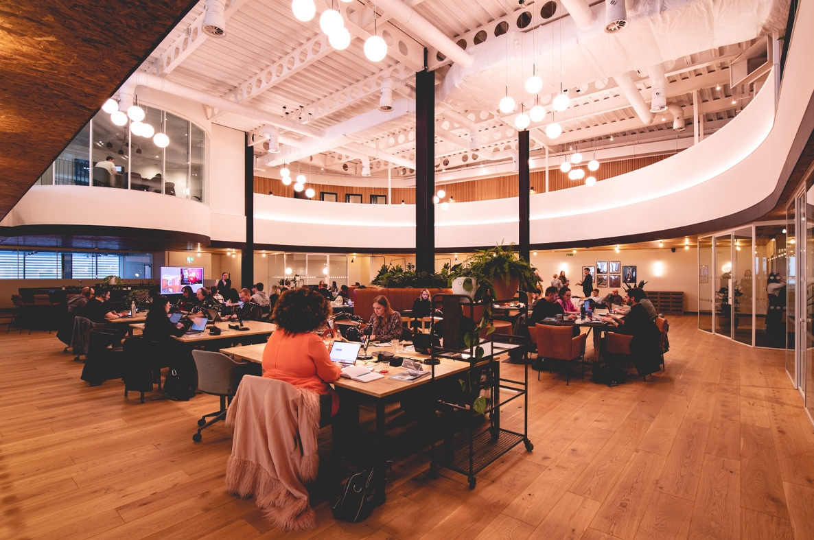 People working at desks in a modern, open-plan office with high ceilings and warm lighting.