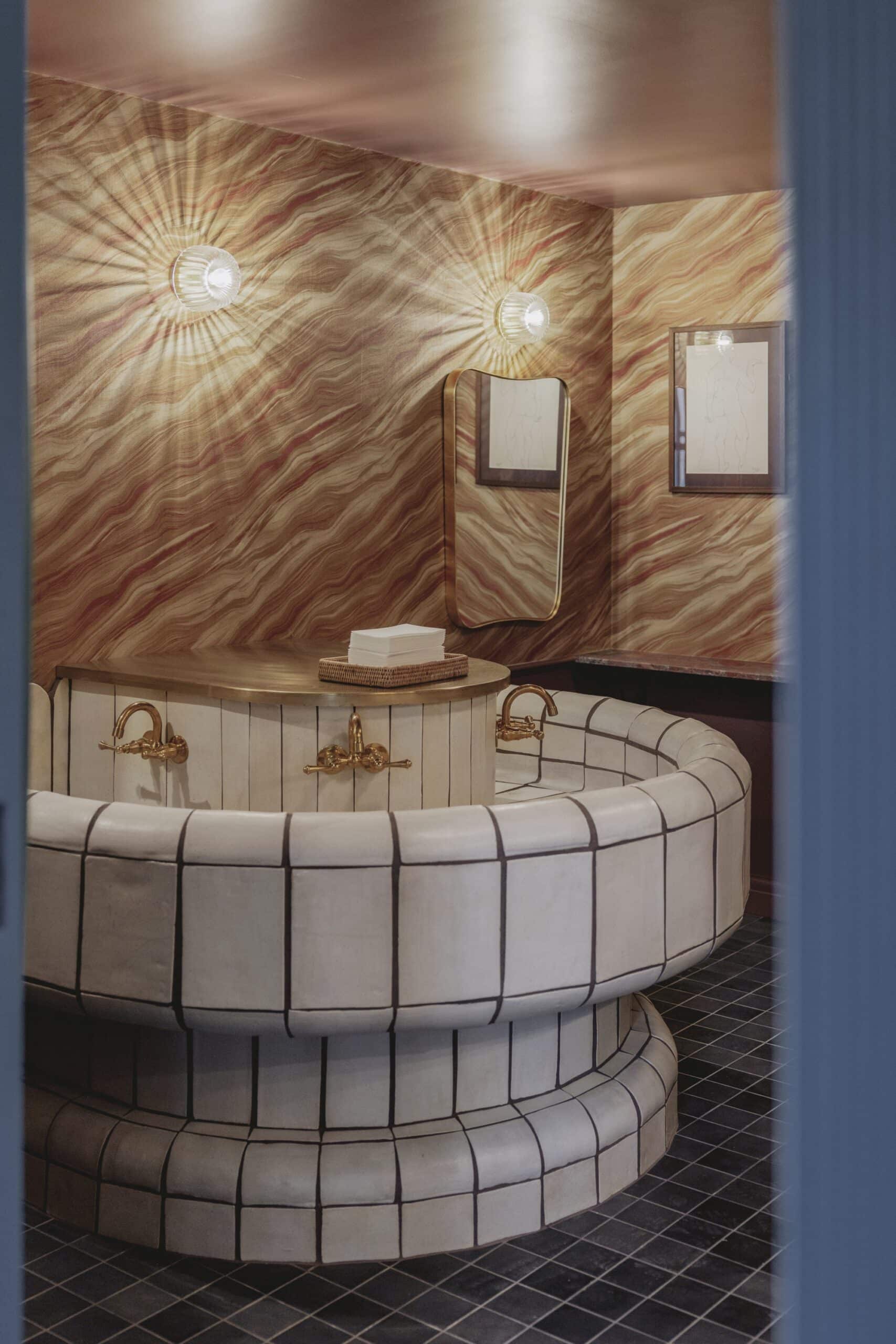 Round tiled wash station with gold faucets and patterned walls under two wall lights.