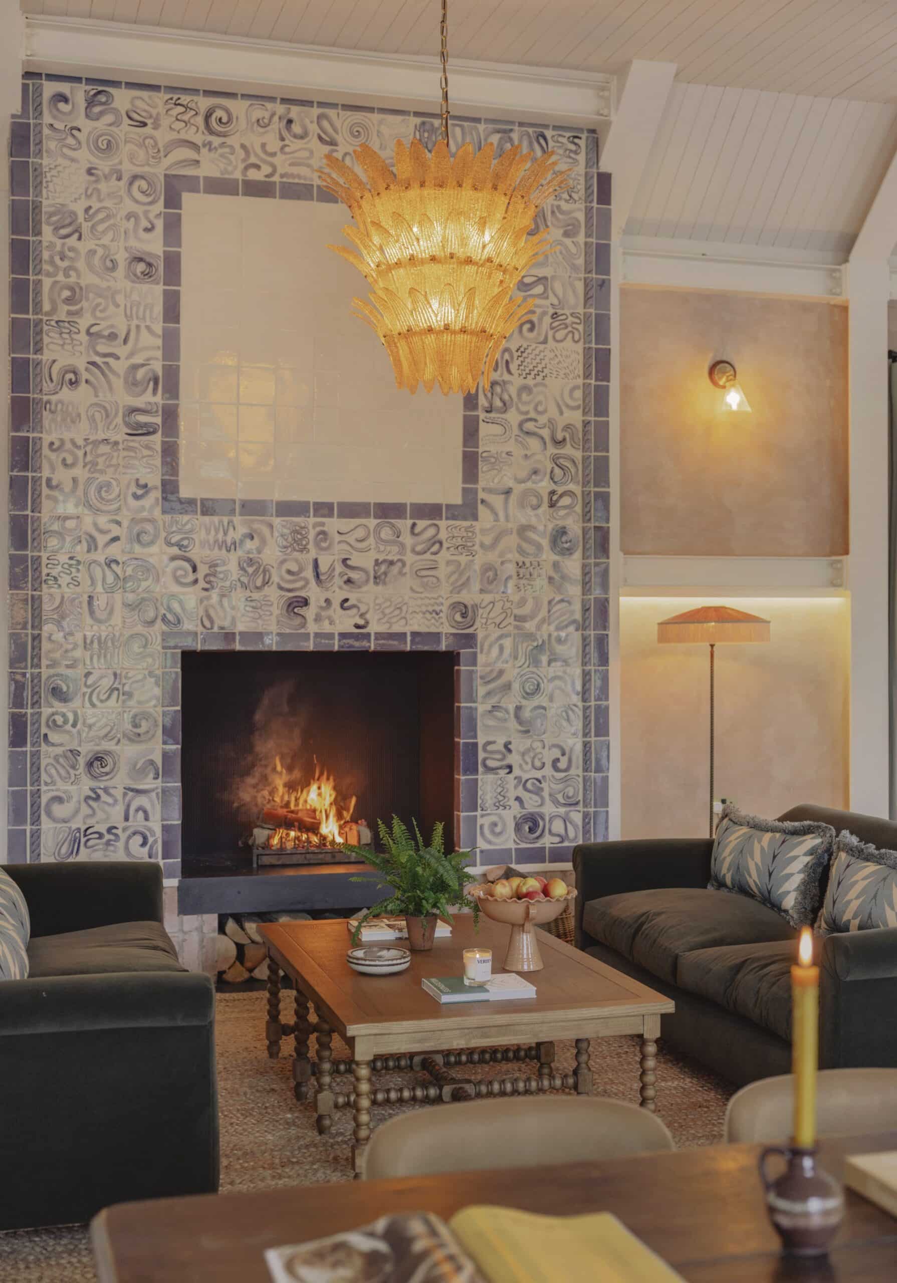 A cozy living room with a lit fireplace, tiled wall, sofas, and a decorative light fixture.
