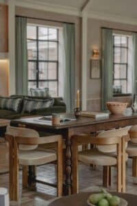 A wooden dining table with chairs, a candle, and a bowl in a cozy, light-filled living room.