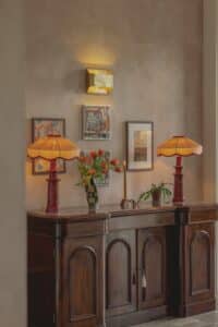 Two orange lamps, flowers, and art on a wooden sideboard against a beige wall.