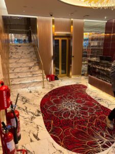 Marble floor lobby with a red carpet, stairs, elevator, and fire extinguishers on the left.