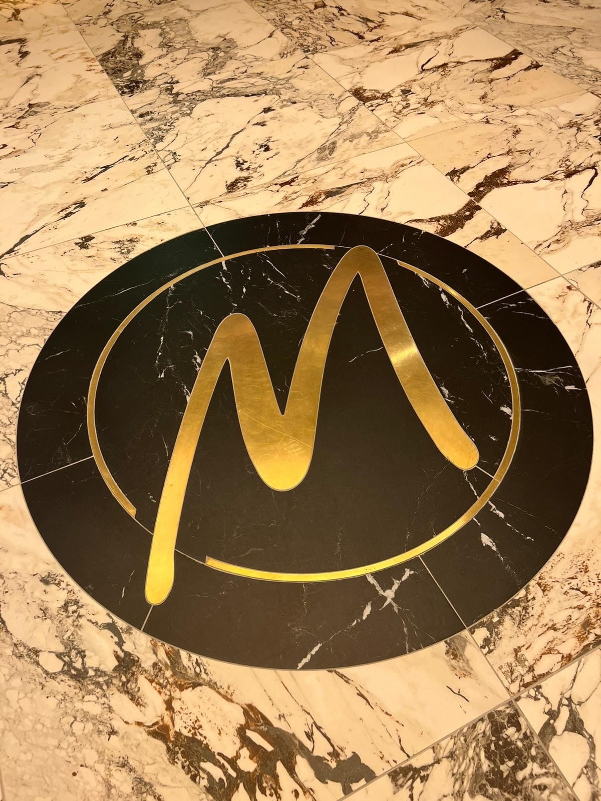 A gold letter "M" in a circle on a marble floor.