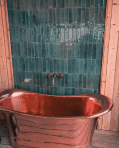 A shiny copper bathtub in front of green tiles and wooden walls.