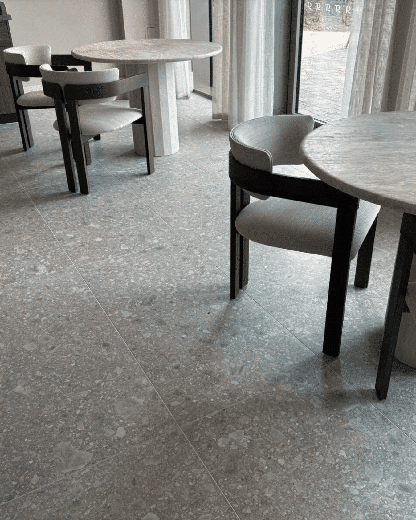 Round tables with modern chairs on a gray tile floor near large windows.