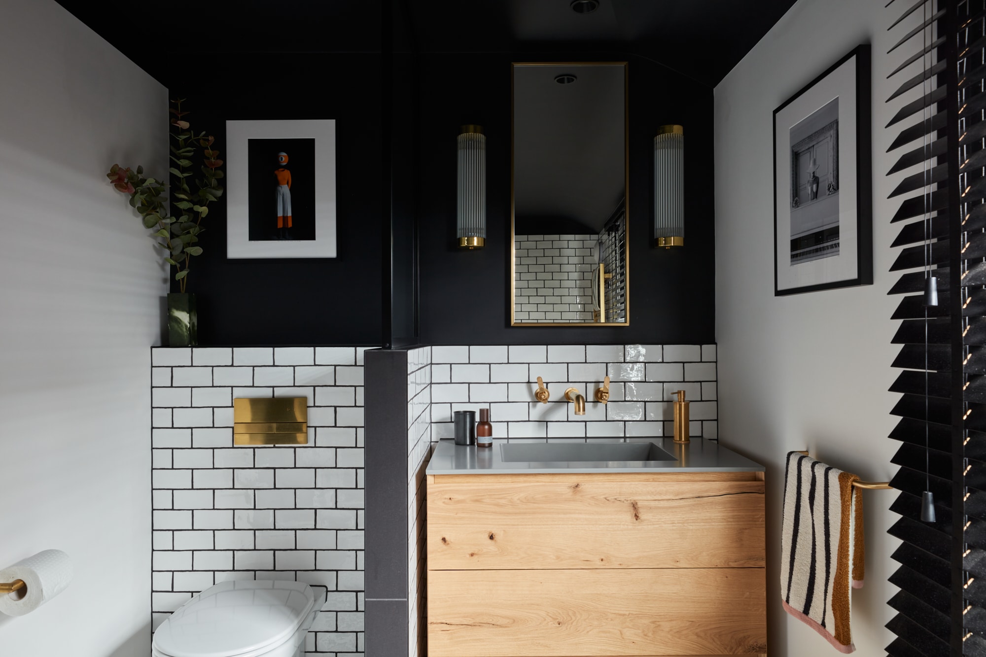 Modern bathroom with white subway tiles, wooden vanity, gold fixtures, and framed art on dark walls.