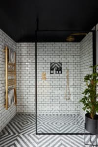 Modern shower with white subway tiles, patterned floor, gold fixtures, and a potted plant in the corner.